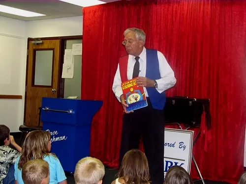 A guest storyteller in a vest presents a book to children gathered in the library meeting room
